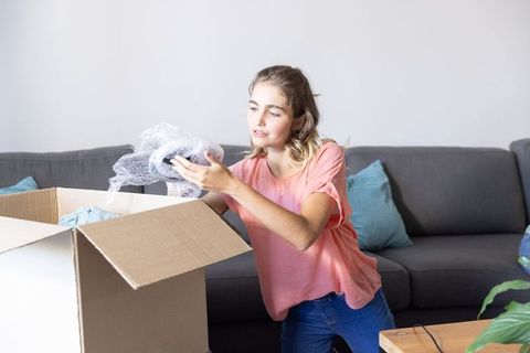 Young woman unpacking cardboard box in living room, inspecting bubble wrap and items