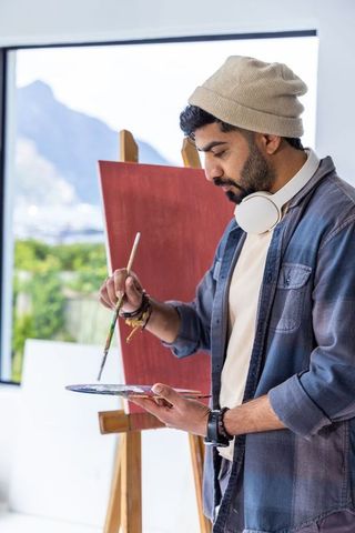 Indian painter painting on red-primed canvas in sunlit studio with mountain view