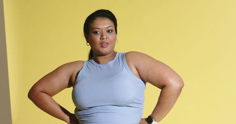 Mid-adult asian woman standing hands on hips in light blue tank top on yellow background