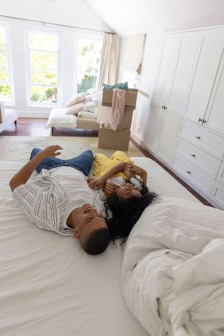 African american couple lying on bed in sunlit new home with moving boxes