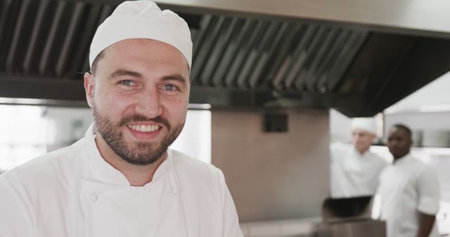 Smiling chef in a professional kitchen, with colleagues in the background. Ideal for advertisements, culinary blogs, restaurant websites, and promotional materials showcasing teamwork, culinary skills, and the happiness of staff in a commercial kitchen environment.