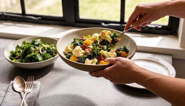 Hands serving roasted cauliflower and kale bowl by sunlit window for healthy meal