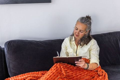Middle-aged woman writing journal on dark gray sofa wrapped in cozy orange blanket