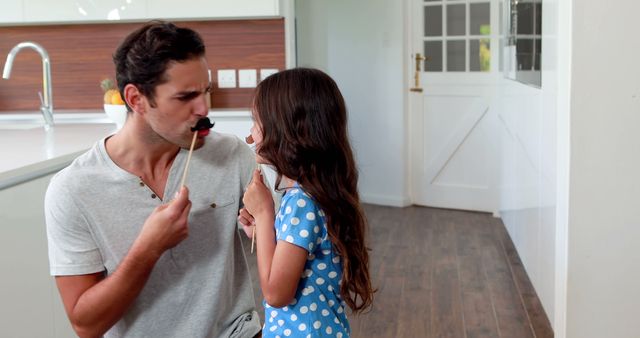 Father and young daughter playing with fake moustaches, enjoying a playful moment together in a cozy home environment. Perfect for illustrating family bonding, father-daughter relationships, fun activities at home, and indoor playtime.