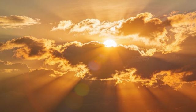 Sun peeking through clouds casting golden crepuscular rays and lens flare across warm sky