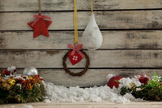 Rustic Christmas decorations featuring a hanging glass lamp, red star, and small wreath against a wooden background. Snow and festive ornaments add to the holiday atmosphere. Ideal for holiday greeting cards, festive invitations, seasonal advertisements, and Christmas-themed blog posts.