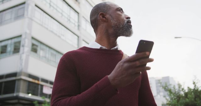 Depicts middle-aged man holding smartphone, standing in urban environment. Useful for promoting mobile technology, urban lifestyle topics, business travel, ICT services, and modern communication advertisement.