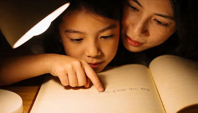 Mother guiding daughter during bedtime reading by warm lamplight