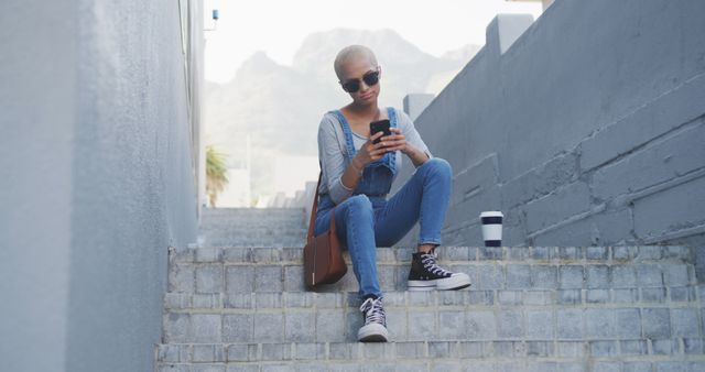 Fashionable biracial woman in sunglasses sitting on steps in city with coffee using smartphone. Summer alternative modern urban lifestyle and communication.