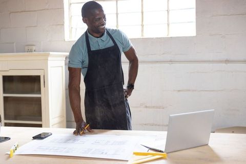 Carpenter reviewing blueprints at workshop table with laptop, pencil and level