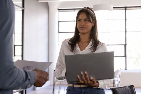 Young professional woman holding laptop discussing project with coworker in bright modern office
