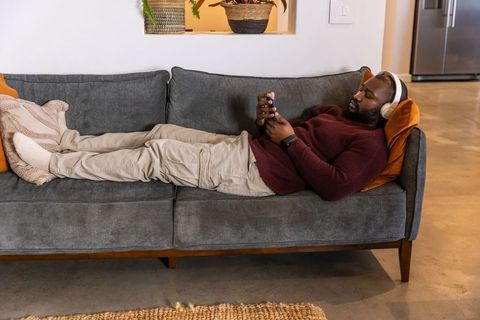 African american man reclining on gray sofa listening to headphones and scrolling smartphone