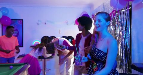 Diverse friends celebrating birthday party with neon lights, pool table and sash