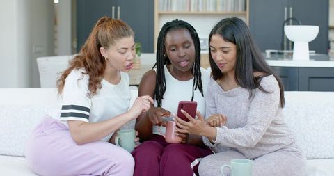 Three friends in loungewear sharing smartphone and coffee while relaxing on pastel sofa