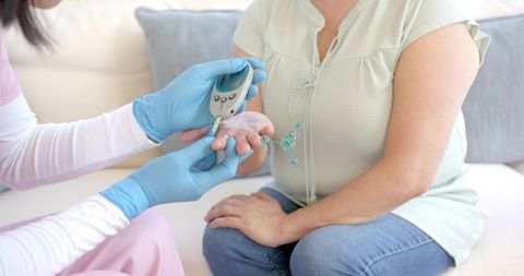 Home nurse testing blood glucose on middle-aged woman using portable glucometer