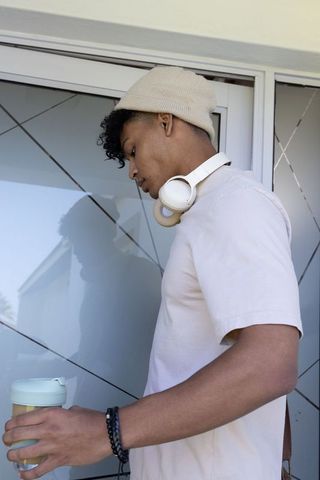 African american man wearing beanie and headphones holding travel mug by glass door