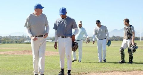 Multigenerational Baseball Team Walking on Sunlit Field Holding Gloves and Catcher Gear