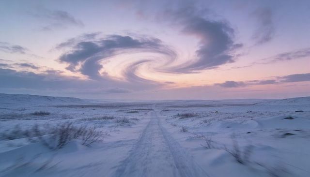 Narrow snow track stretching toward horizon beneath spiral cloud at pastel dawn