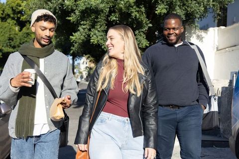 Diverse friends walking and laughing on sunlit city street with coffee and bags