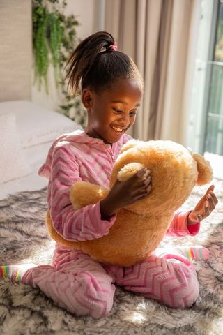 Smiling girl hugging large teddy bear in pink pajamas by sunlit bedroom window