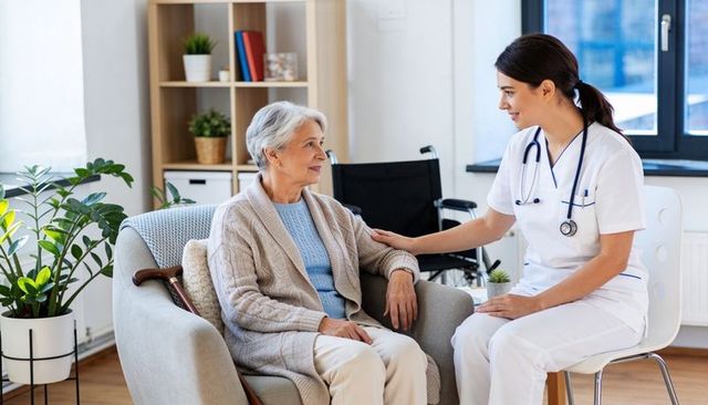 Compassionate nurse supporting elderly woman at home, wheelchair, cane, stethoscope
