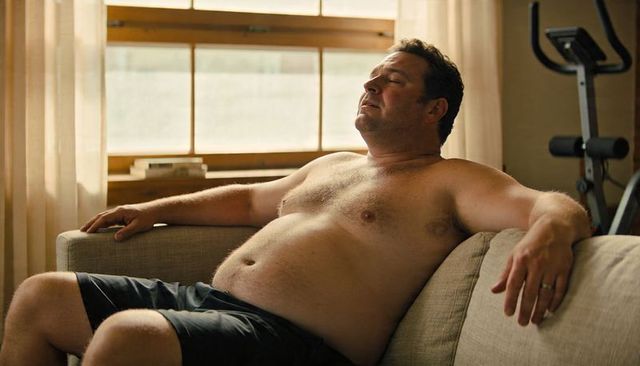 Shirtless middle-aged man reclining on sofa in sunlit living room with exercise bike