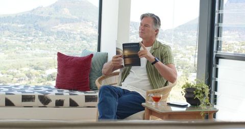 Senior man reading paperback in wicker chair by sunlit window overlooking valley view