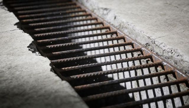 Rusted metal grate running diagonally across cracked concrete curb casting linear shadows