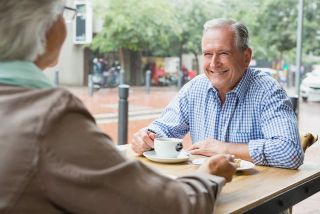 Senior couple sitting at an outdoor cafe, enjoying coffee and engaging in conversation. Ideal for use in advertisements for retirement communities, coffee shops, or lifestyle blogs focusing on senior living and relationships.