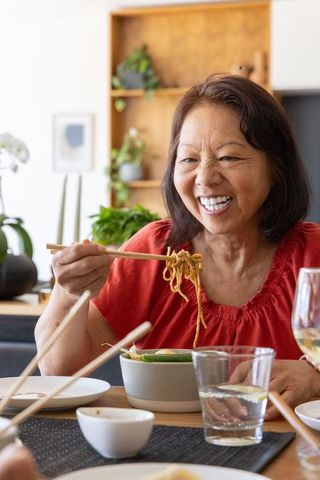 Senior asian woman smiling while eating noodles with chopsticks in cozy home dining