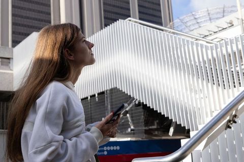 Young woman holding smartphone looking up at modern white slatted staircase urban plaza