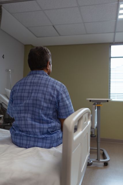 Rear view of mature male patient sitting on medical bed and looking through window in medical ward at hospital