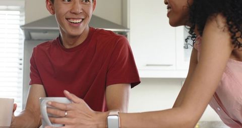 Asian man and african american woman sharing morning coffee in bright kitchen, checking smartwatch