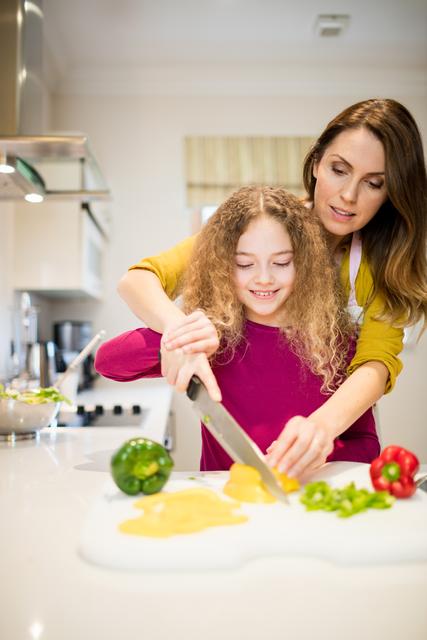 Mother assisting daughter in cutting vegetables in kitchen at home
