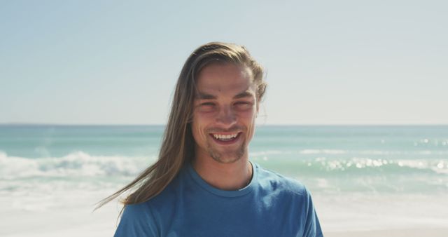 Young man with long hair enjoying a sunny day on the beach while smiling. Ideal for concepts of vacation, summer fun, relaxation, and carefree lifestyle. Perfect for travel brochures, beachside resort advertising, posters, websites, and blog content focused on leisure activities.