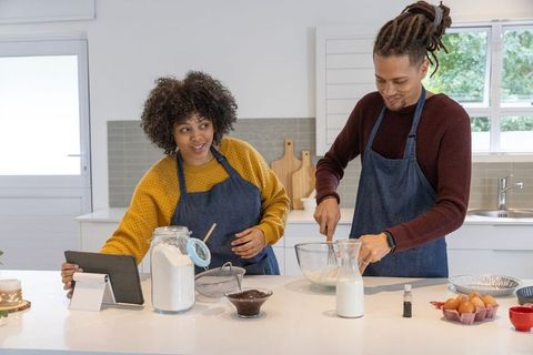 Couple baking together in modern kitchen using tablet for recipe, whisking batter, smiling