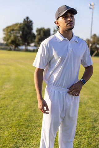 Indian athlete standing in white cricket uniform and cap on sunlit grass field