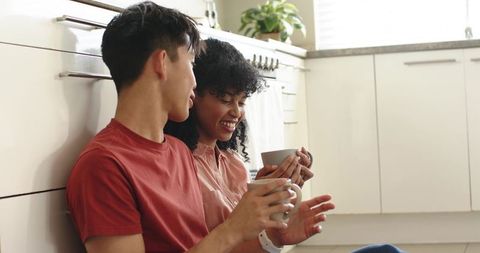 Cozy diverse couple sipping coffee on kitchen floor enjoying casual morning together