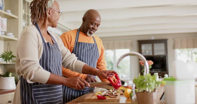Senior African-American couple are enjoying cooking together in a modern, brightly lit kitchen. The woman is slicing a red bell pepper, and the man is chopping mushrooms. Their expressions show joy and companionship. This image can be used to promote senior lifestyles, healthy home-cooked meals, family bonding activities, and retirement living communities.