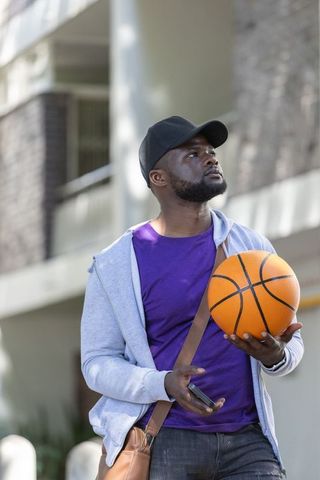 Young black man holding basketball and smartphone standing by urban apartment building