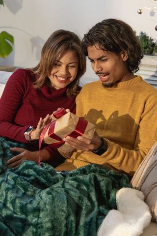 Couple unwrapping gift on couch under green plush blanket sharing cozy romantic holiday