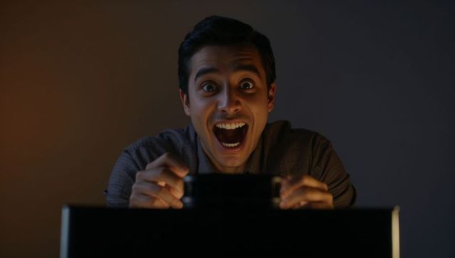 Excited man reacting to smartphone over monitor, celebrating big win in dark room