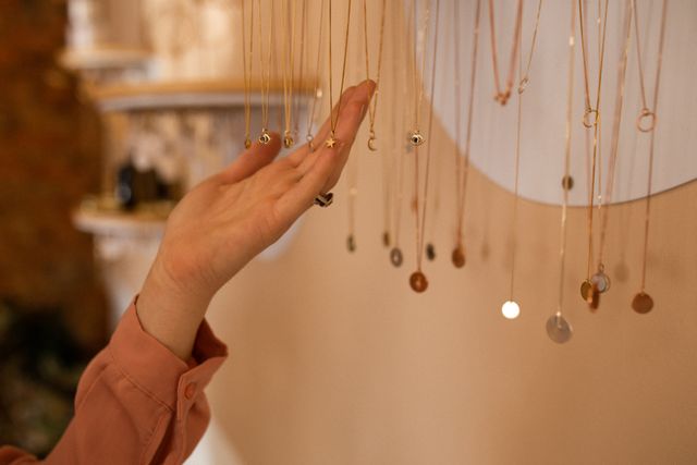 Hand of a caucasian female customer touching delicate necklaces hanging on display in a jewellery shop. Ideal for use in articles or advertisements about independent handmade craft businesses, shopping experiences, and fashion accessories.