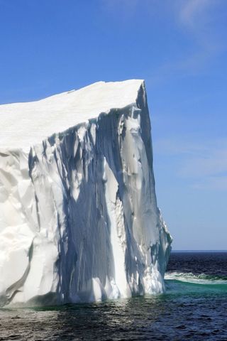 Towering iceberg cliff rising from deep blue ocean with sculpted ice formations under clear sky