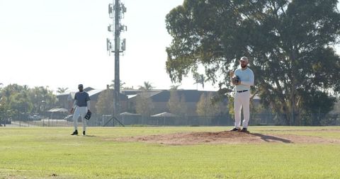 Pitcher Standing on Mound with Teammate Waiting on Sunlit Baseball Field