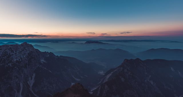 Serene mountain ranges are seen during the twilight hour, with layers of peaks and valleys fading into the horizon. Ideal for projects related to nature, travel, adventure, and outdoor activities. Perfect for use in wallpapers, promotional materials, and backgrounds emphasizing tranquility and natural beauty.