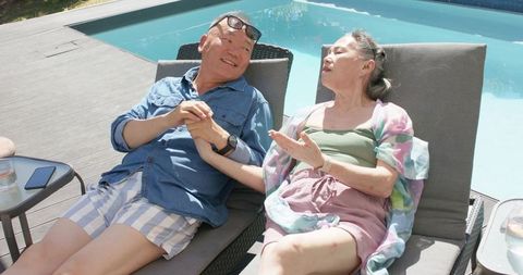 Senior asian couple relaxing by pool holding hands on sunlit deck, summer romance