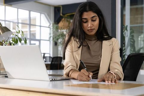 Indian businesswoman writing at desk in modern coworking office with laptop and plants