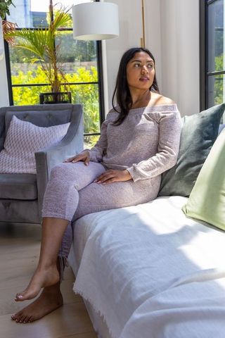 Sunlit african american woman sitting on daybed in knit loungewear, barefoot, cozy interior
