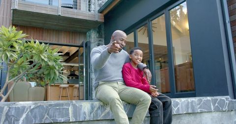 Smiling african american father pointing while sitting with son on modern patio